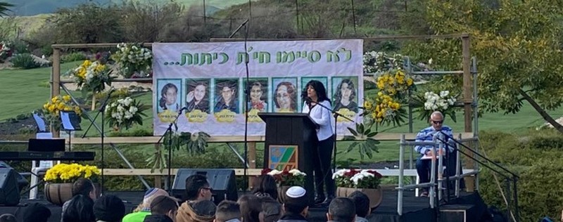 במה ועליה נואמת. מאחור - תמונה של שבע הבנות שנרצחו